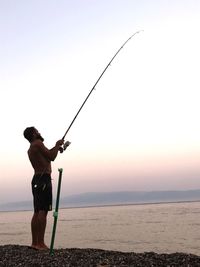 Side view of shirtless man fishing in sea against clear sky during sunset