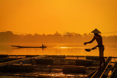 Silhouette fisherman feeding against orange sky