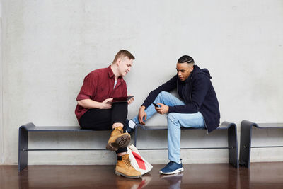 Full length of young male students using technologies while sitting on bench against wall in university