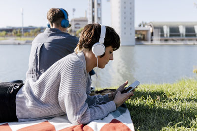 Woman wearing headphones using mobile phone while lying on blanket by friend against river