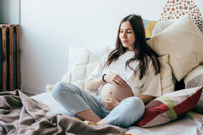 Young caucasian pregnant woman is sitting on the bed in the apartment and touching her belly