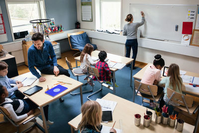 High angle view of female teacher writing on board while male educator sitting with students at desk in classroom