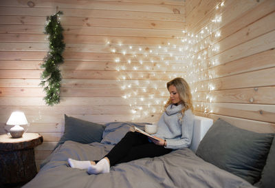 Young woman reading book while sitting on bed at home during christmas