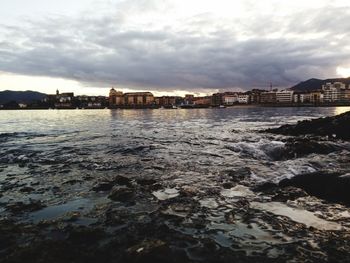 View of city at waterfront against cloudy sky
