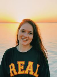 Portrait of young woman standing at beach against sky during sunset