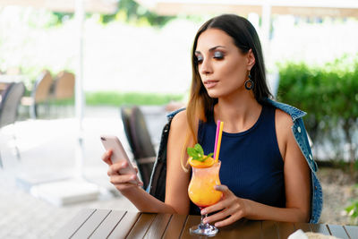 Young woman with drink using smart phone while sitting at sidewalk cafe