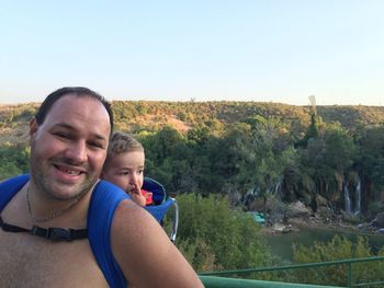 Portrait of happy man carrying son in baby carrier against forest