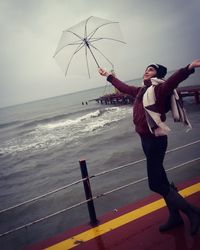 Carefree woman holding umbrella on pier over sea against cloudy sky