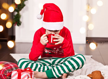Boy kid in funny red santa cap and christmas pajamas sitting on a plaid and drinking cocoa