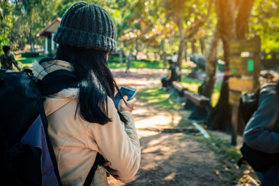 Side view of young woman with backpack using mobile phone while standing at park
