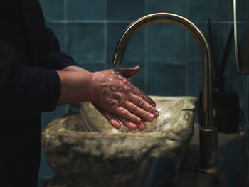 Elderly senior lady in black clothes washes her hands under the tap in the stone washbasin .