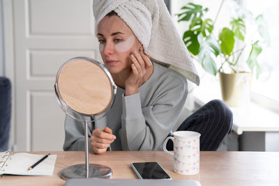 Woman with towel on head applying patches under eyes, looking at mirror. face skin care beauty 