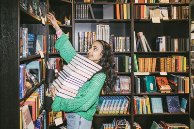 Girl with heap of books reaching at shelf while standing in bookstore