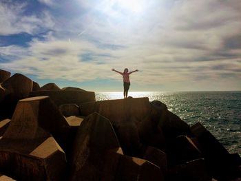 Young woman with arms outstretched standing on rock by sea against sky