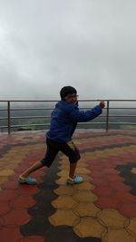 Full length portrait of smiling boy posing at observation point against cloudy sky