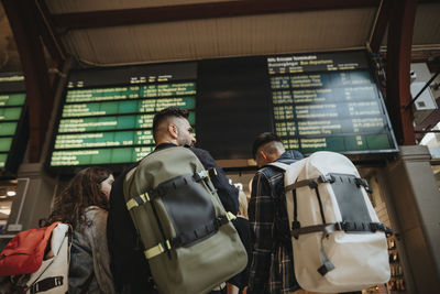 Rear view of family with backpacks standing against arrival departure board at railroad station