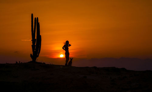 Silhouette woman standing with dog on field against orange sky during sunset