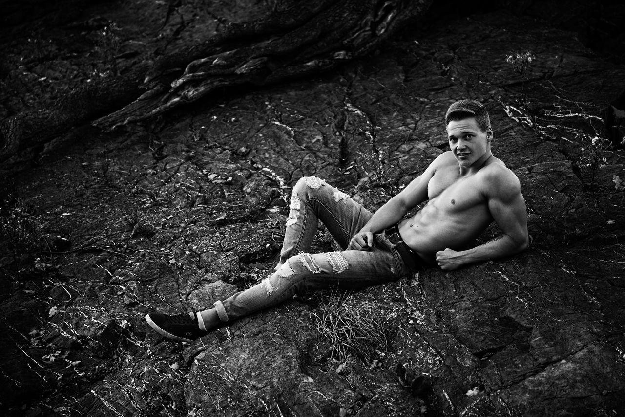 real people, one person, lifestyles, leisure activity, full length, rock, shirtless, land, rock - object, nature, solid, young men, young adult, sitting, day, forest, tree, looking away, outdoors, shorts