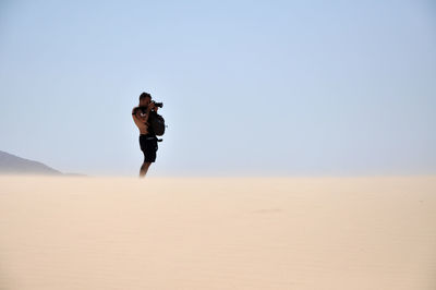 Young man photographing while standing in desert against clear sky