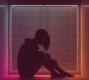 Side view of woman sitting by window blinds at home