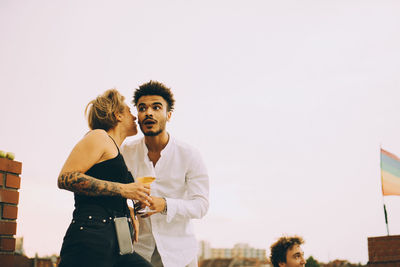 Woman having drink while sharing gossip with young man on terrace at rooftop party