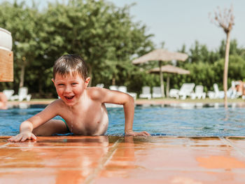 Delighted cute child with wet hair leaning on poolside and looking at camera while having fun during summer weekend