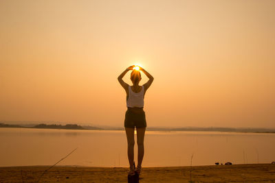 Rear view of woman gesturing while standing at lake against sky during sunset