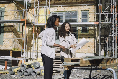 Female design professional showing blueprints to client while standing in front of under construction apartment