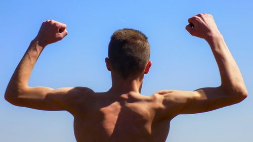 Rear view of shirtless man flexing muscles against clear sky