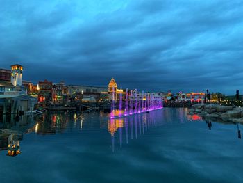 Illuminated buildings by river against cloudy sky