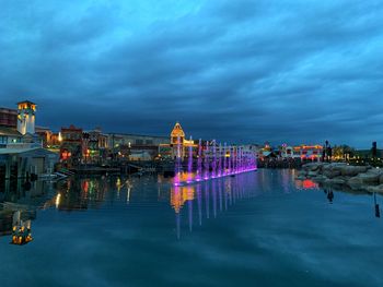 Illuminated buildings by river against cloudy sky