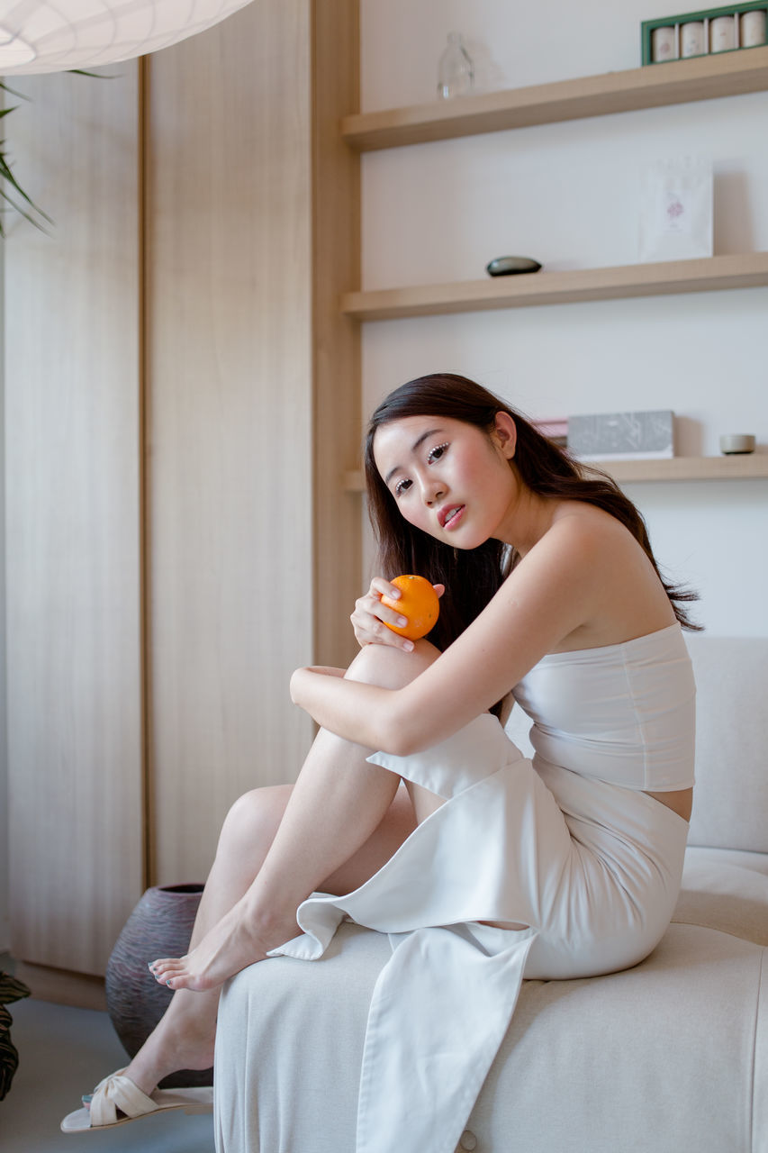 FULL LENGTH OF A SMILING YOUNG WOMAN SITTING ON WALL