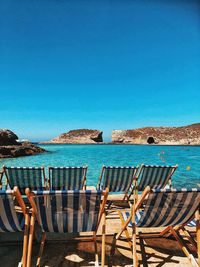 Empty chairs by sea against clear blue sky