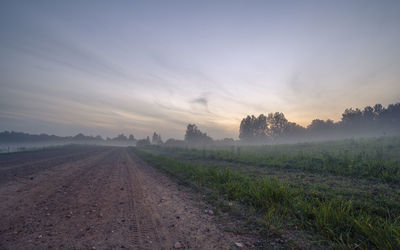 Dirt road amidst field against sky during sunset