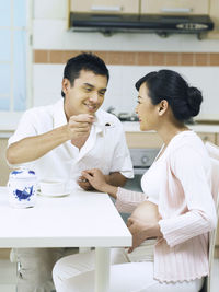 Man feeding pregnant wife while sitting at table in home