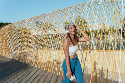 Contemplative young ethnic female in bandana and wireless headset listening to song while closed eyes on urban bridge