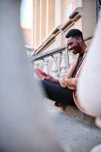 Modern cheerful black african american man wearing stylish clothes and earring browsing on tablet while sitting in stone stairway on the street