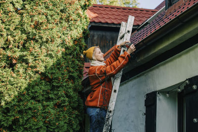 Smiling woman standing on ladder decorating house with lights for christmas