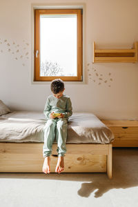Full body of boy in sleepwear playing game on cellphone while sitting on bed near window in light bedroom
