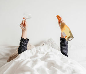 Person in bed, covered with white linen, holding champagne bottle and glass.