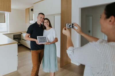Woman photographing smiling couple with digital tablet standing at home