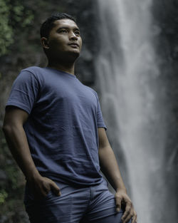 Young man looking away standing against waterfall in forest