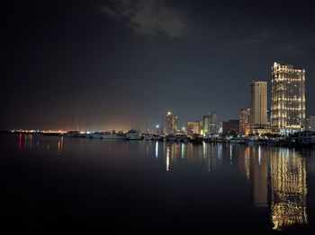 Illuminated buildings by sea against sky at night