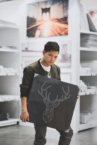 Portrait of man holding drawing on blackboard while standing on floor