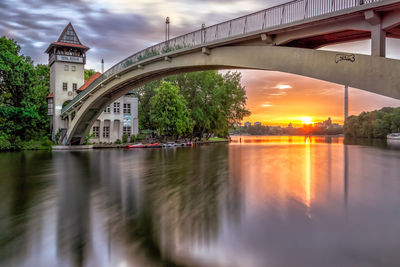 Arch bridge over river by buildings against sky during sunset