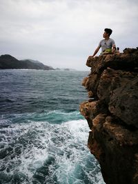 Young man sitting on cliff by sea against sky