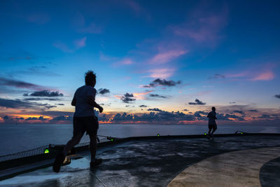 Silhouette people running on walkway by sea at sunset