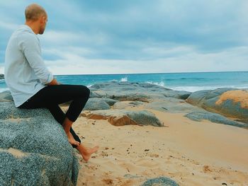 Man sitting on rock at beach against sky