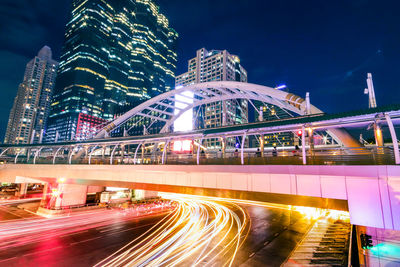 Light trails on bridge by buildings against sky at night