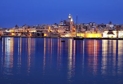 Illuminated buildings by river against blue sky